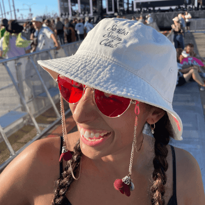 Girl wearing unstructured ivory bucket hat with grey script embroidery Santos Swim Club and Roseys rose colored classes with sunglass chains at music festival